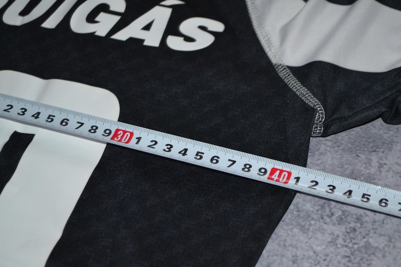 Puede incluir: Una camiseta gris oscuro con el texto blanco "QUIG&Aacute;S". Una regla muestra una medida de 30 a 40 cm. La camiseta tiene mangas a rayas blancas y grises. La tela parece ser un material de punto suave.