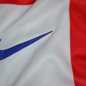 Puede incluir: Una prenda de tela blanca y roja con un logotipo Nike swoosh azul bordado. El material muestra una textura suave con pliegues y arrugas visibles, resaltando el contraste entre los paneles de color rojo brillante y las secciones blancas.