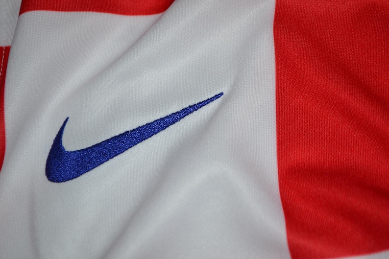 Puede incluir: Una prenda de tela blanca y roja con un logotipo Nike swoosh azul bordado. El material muestra una textura suave con pliegues y arrugas visibles, resaltando el contraste entre los paneles de color rojo brillante y las secciones blancas.