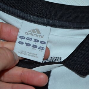 May include: Close-up of a white and black Adidas jersey. The jersey has a black collar and sleeve cuffs. A tag with the Adidas logo and size information is visible. The tag reads "Made in Tunisia / Fabriqu&eacute; en Tunisie."