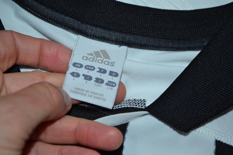 May include: Close-up of a white and black Adidas jersey. The jersey has a black collar and sleeve cuffs. A tag with the Adidas logo and size information is visible. The tag reads "Made in Tunisia / Fabriqu&eacute; en Tunisie."