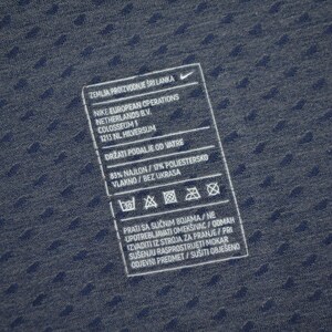 May include: Close-up of a navy blue fabric with a textured pattern. A white rectangular tag with black text is visible, including the Nike logo and care instructions. The tag provides details about the fabric composition and washing guidelines.