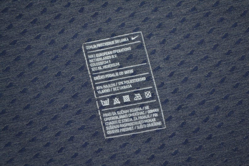 May include: Close-up of a navy blue fabric with a textured pattern. A white rectangular tag with black text is visible, including the Nike logo and care instructions. The tag provides details about the fabric composition and washing guidelines.
