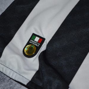 Puede incluir: Primer plano de una tela a rayas blancas y negras, posiblemente una camiseta o camisa. Un peque&ntilde;o emblema con la bandera italiana y la inscripci&oacute;n "PRODOTTO UFFICIALE" est&aacute; adjunto. El emblema tiene un elemento circular dorado.