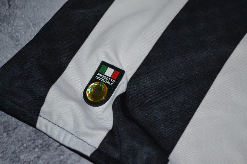 Puede incluir: Primer plano de una tela a rayas blancas y negras, posiblemente una camiseta o camisa. Un peque&ntilde;o emblema con la bandera italiana y la inscripci&oacute;n "PRODOTTO UFFICIALE" est&aacute; adjunto. El emblema tiene un elemento circular dorado.