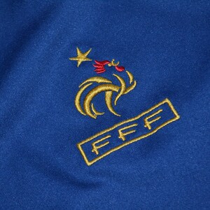 May include: Close-up of blue fabric with gold embroidery. The embroidery depicts a star, a rooster, and the letters "FFF" within a rectangle. The design is likely a sports emblem or logo.
