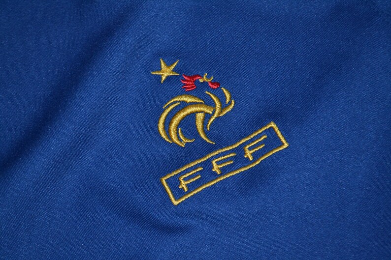 May include: Close-up of blue fabric with gold embroidery. The embroidery depicts a star, a rooster, and the letters "FFF" within a rectangle. The design is likely a sports emblem or logo.