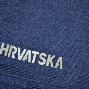 May include: Close-up of a navy blue fabric with the word "HRVATSKA" printed in silver. The text is slightly angled and appears to be part of a sports uniform or athletic wear.