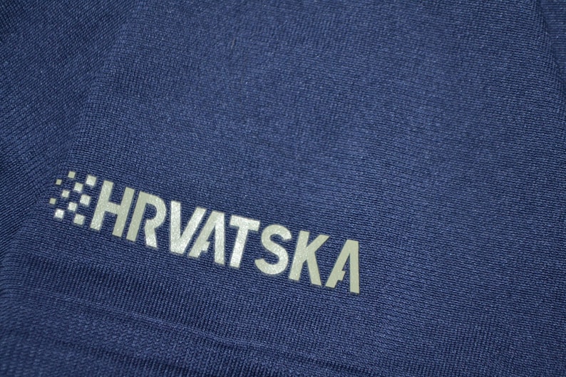 May include: Close-up of a navy blue fabric with the word "HRVATSKA" printed in silver. The text is slightly angled and appears to be part of a sports uniform or athletic wear.