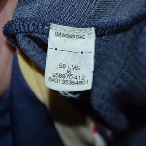 May include: Close-up of a clothing tag with the text "IM#363545", "S8 LMS XL", and several number sequences. The tag is attached to a dark blue fabric, with a yellow and red fabric visible in the background.
