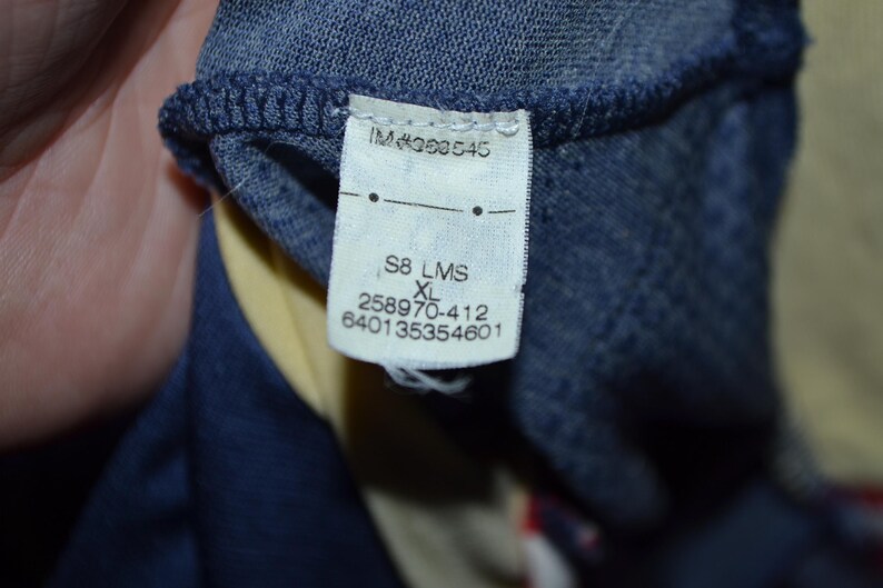 May include: Close-up of a clothing tag with the text "IM#363545", "S8 LMS XL", and several number sequences. The tag is attached to a dark blue fabric, with a yellow and red fabric visible in the background.