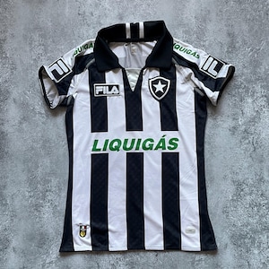 Puede incluir: Una camiseta de f&uacute;tbol a rayas blancas y negras con cuello y mangas negras. La camiseta presenta la marca "FILA" y la palabra "LIQUIG&Aacute;S" en verde. Un escudo de estrella est&aacute; en el pecho.