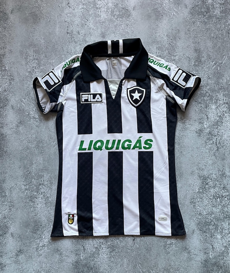 Puede incluir: Una camiseta de f&uacute;tbol a rayas blancas y negras con cuello y mangas negras. La camiseta presenta la marca "FILA" y la palabra "LIQUIG&Aacute;S" en verde. Un escudo de estrella est&aacute; en el pecho.