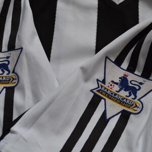 May include: A close-up of a black and white striped football shirt. The sleeves display the Barclays Premier League logo, featuring a blue lion, a football, and the words "Barclaycard Premiership".