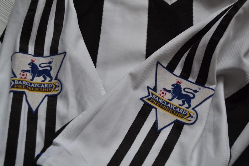 May include: A close-up of a black and white striped football shirt. The sleeves display the Barclays Premier League logo, featuring a blue lion, a football, and the words "Barclaycard Premiership".