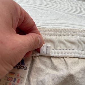 Puede incluir: Primer plano de una etiqueta de prenda Adidas. La etiqueta muestra el logotipo de Adidas y varias banderas internacionales. La prenda es de color beige claro. La etiqueta tambi&eacute;n tiene los n&uacute;meros 627117 y ASR001/02.