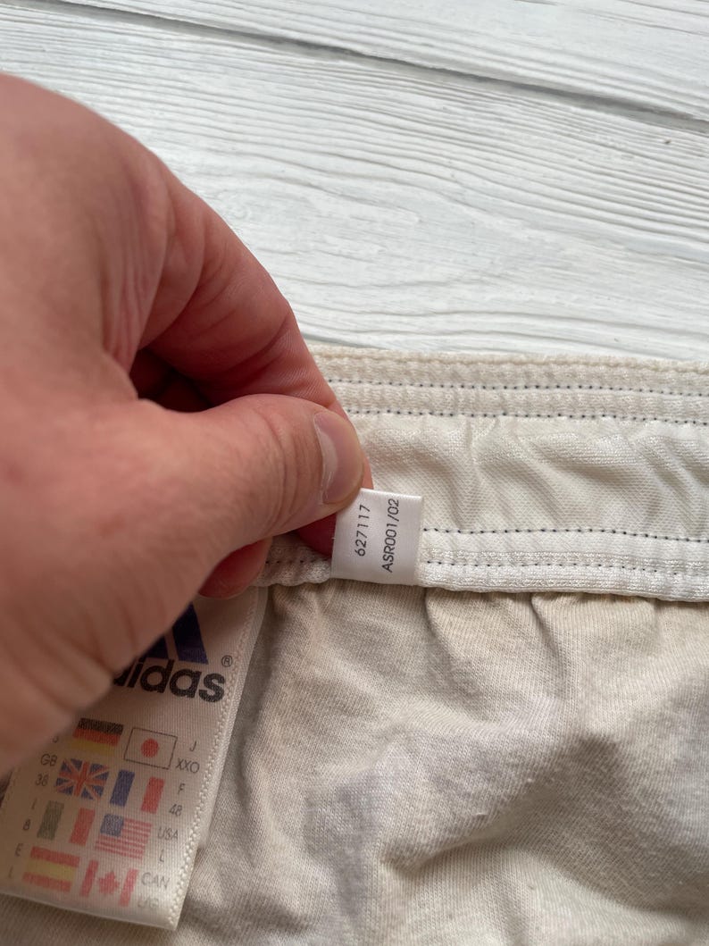 Puede incluir: Primer plano de una etiqueta de prenda Adidas. La etiqueta muestra el logotipo de Adidas y varias banderas internacionales. La prenda es de color beige claro. La etiqueta tambi&eacute;n tiene los n&uacute;meros 627117 y ASR001/02.