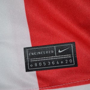 Puede incluir: Un panel de tela rojo y blanco con una etiqueta rectangular negra que contiene el texto blanco ENGINEERED, un logotipo de Nike swoosh y el c&oacute;digo alfanum&eacute;rico o 805364n 20. La etiqueta est&aacute; cosida al material con hilo blanco visible.