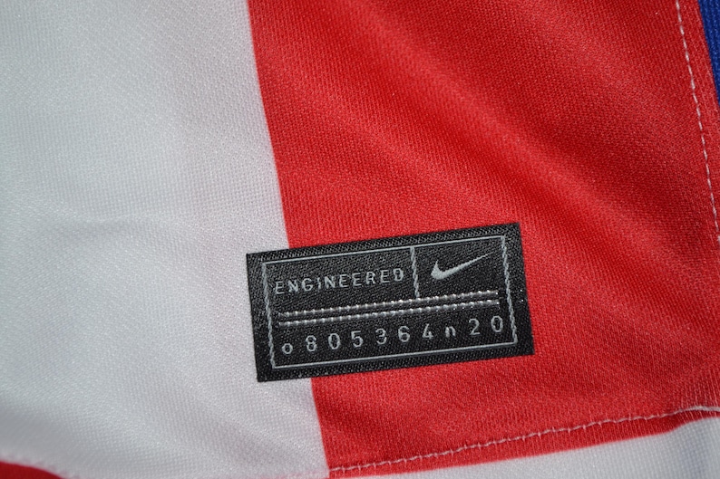 Puede incluir: Un panel de tela rojo y blanco con una etiqueta rectangular negra que contiene el texto blanco ENGINEERED, un logotipo de Nike swoosh y el c&oacute;digo alfanum&eacute;rico o 805364n 20. La etiqueta est&aacute; cosida al material con hilo blanco visible.
