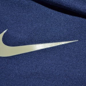 May include: Close-up of navy blue fabric with a large, white Nike swoosh logo. The logo is positioned diagonally across the frame, with the fabric's texture visible. The image highlights the brand's iconic symbol.