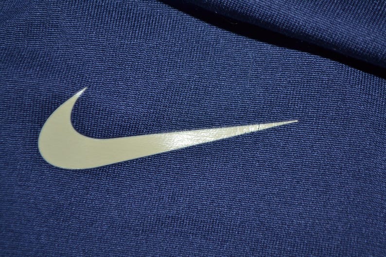 May include: Close-up of navy blue fabric with a large, white Nike swoosh logo. The logo is positioned diagonally across the frame, with the fabric's texture visible. The image highlights the brand's iconic symbol.