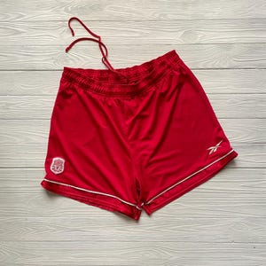 May include: Bright red athletic shorts with a white trim and drawstring. The shorts feature a team logo on the left leg and the Reebok logo on the right leg. The shorts are made of a lightweight, breathable material.