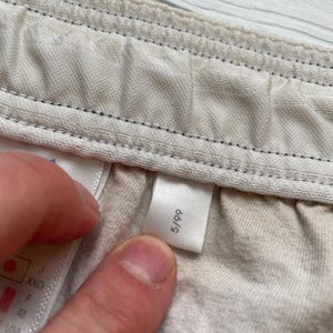 Puede incluir: Primer plano de una etiqueta con el n&uacute;mero 5/99 impreso, adherida a una tela de color claro. La tela parece ser parte de una prenda, con costuras visibles y una superficie texturizada.
