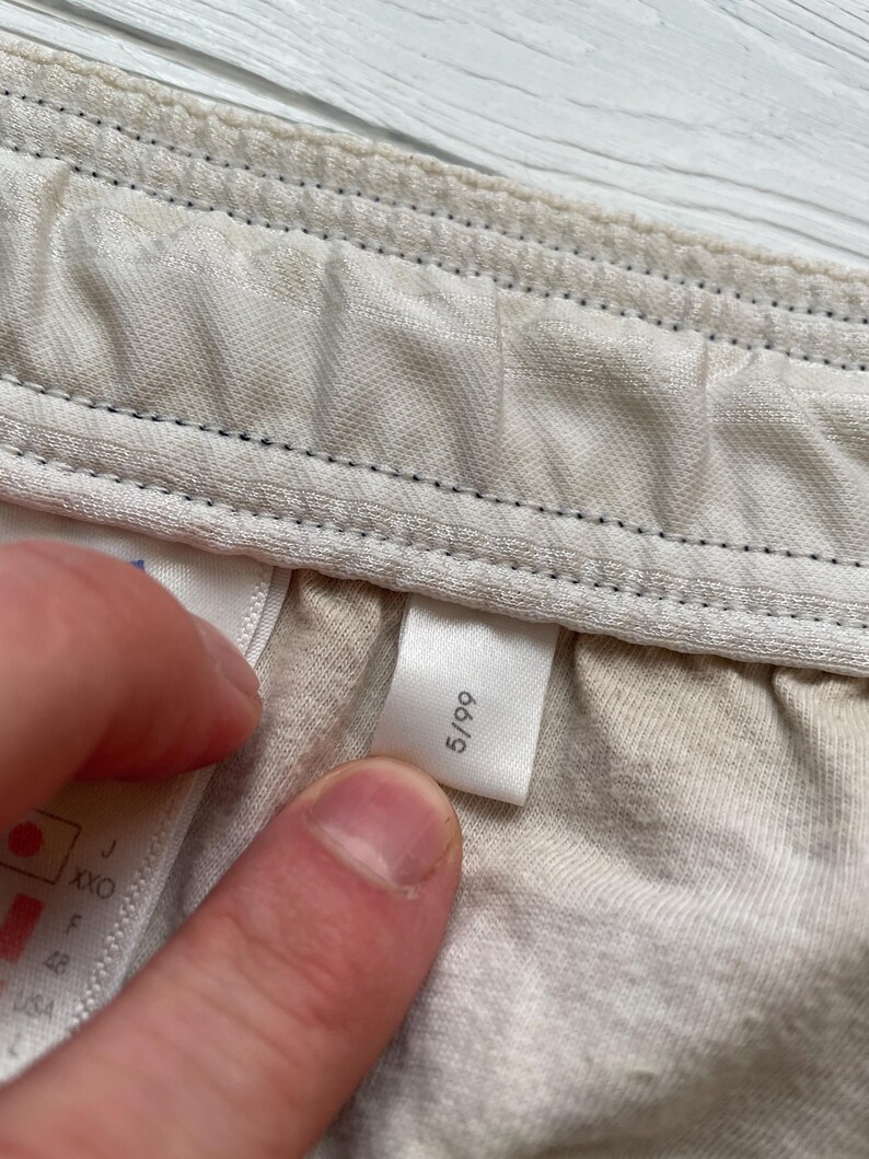 Puede incluir: Primer plano de una etiqueta con el n&uacute;mero 5/99 impreso, adherida a una tela de color claro. La tela parece ser parte de una prenda, con costuras visibles y una superficie texturizada.