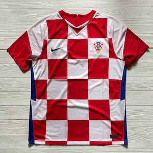 Puede incluir: Una camiseta de f&uacute;tbol de manga corta con un patr&oacute;n de cuadros rojos y blancos en la parte delantera. La prenda incluye un logotipo Nike azul marino en el pecho derecho y el escudo de la Federaci&oacute;n Croata de F&uacute;tbol HNS en el pecho izquierdo.