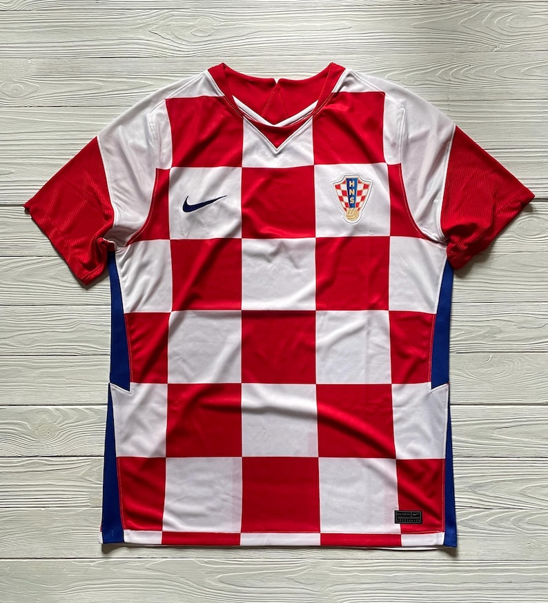 Puede incluir: Una camiseta de f&uacute;tbol de manga corta con un patr&oacute;n de cuadros rojos y blancos en la parte delantera. La prenda incluye un logotipo Nike azul marino en el pecho derecho y el escudo de la Federaci&oacute;n Croata de F&uacute;tbol HNS en el pecho izquierdo.