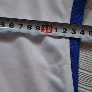 Puede incluir: Una prenda blanca con un borde acanalado azul a lo largo del lateral. Una cinta m&eacute;trica met&aacute;lica est&aacute; colocada horizontalmente sobre la tela, mostrando una medida que supera los 50 cent&iacute;metros, indicando un ancho de unos 57 cent&iacute;metros.