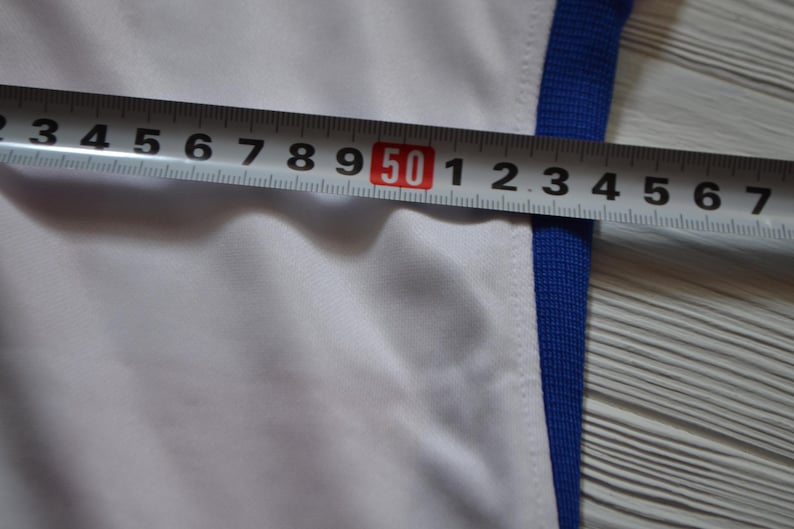 Puede incluir: Una prenda blanca con un borde acanalado azul a lo largo del lateral. Una cinta m&eacute;trica met&aacute;lica est&aacute; colocada horizontalmente sobre la tela, mostrando una medida que supera los 50 cent&iacute;metros, indicando un ancho de unos 57 cent&iacute;metros.