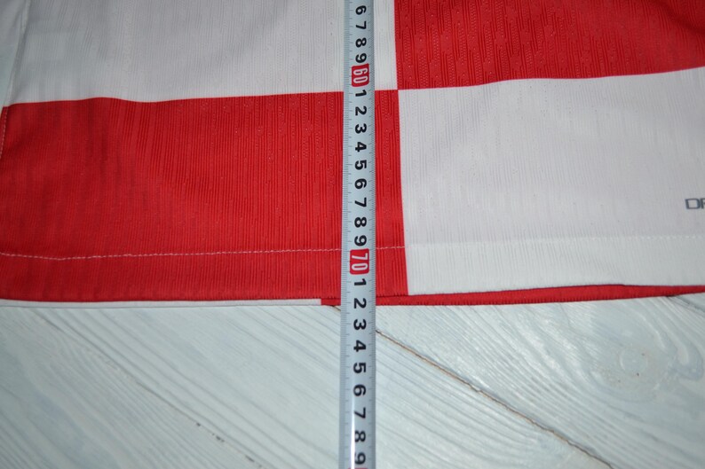 May include: A red and white checkered shirt featuring a visible measuring tape placed vertically against the fabric. The tape indicates a length measurement extending past 72 centimeters, showing the scale of the garment's patterned textile.
