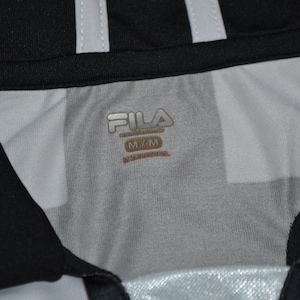 Puede incluir: Primer plano del cuello de una camiseta deportiva en blanco y negro. La camiseta tiene un cuello negro y rayas blancas. El logotipo de Fila es visible en dorado, junto con la talla M/M.