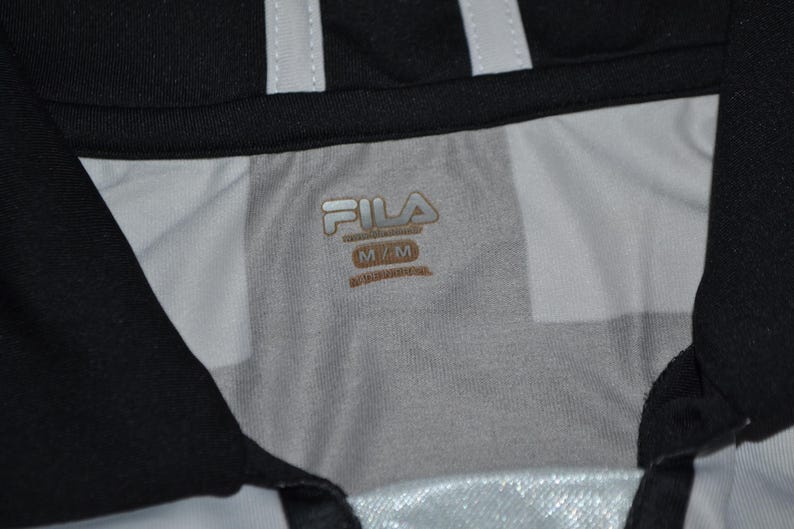 Puede incluir: Primer plano del cuello de una camiseta deportiva en blanco y negro. La camiseta tiene un cuello negro y rayas blancas. El logotipo de Fila es visible en dorado, junto con la talla M/M.