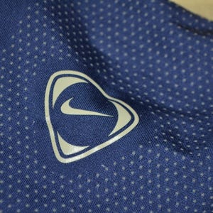 May include: Close-up of a blue athletic shirt with a white Nike logo in a heart-shaped design. The shirt has a dotted pattern across the fabric. The logo is a white swoosh inside a white heart-shaped outline.