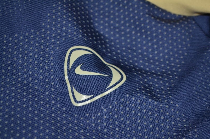 May include: Close-up of a blue athletic shirt with a white Nike logo in a heart-shaped design. The shirt has a dotted pattern across the fabric. The logo is a white swoosh inside a white heart-shaped outline.