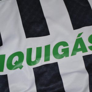 Puede incluir: Primer plano de una tela blanca con rayas verticales negras. La palabra "LIQUIG&Aacute;S" est&aacute; impresa en letras verdes grandes en la tela. La tela parece ser parte de una camiseta o uniforme deportivo.