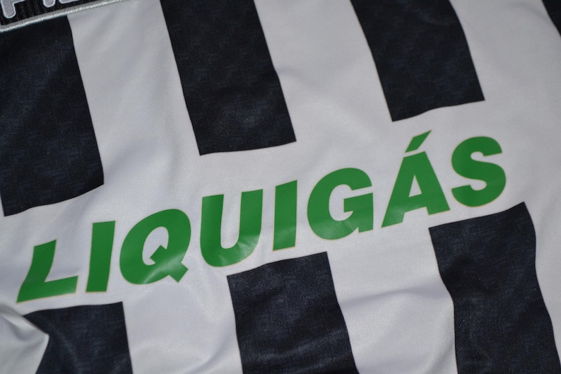 Puede incluir: Primer plano de una tela blanca con rayas verticales negras. La palabra "LIQUIG&Aacute;S" est&aacute; impresa en letras verdes grandes en la tela. La tela parece ser parte de una camiseta o uniforme deportivo.