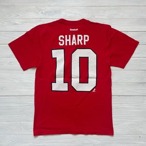 May include: Red t-shirt with the name "SHARP" and the number "10" in white block letters. The Reebok logo is visible at the top. The shirt is short-sleeved and appears to be made of a soft material.