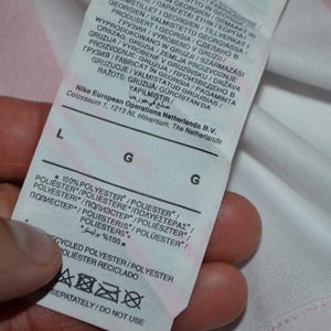 Puede incluir: Una etiqueta de cuidado de tela blanca unida a una prenda, con el texto Nike European Operations Netherlands B.V., Colosseum 1, 1213 NL, Hilversum, The Netherlands. La etiqueta indica una talla L, composici&oacute;n 100% poli&eacute;ster e instrucciones de cuidado.