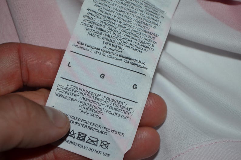 Puede incluir: Una etiqueta de cuidado de tela blanca unida a una prenda, con el texto Nike European Operations Netherlands B.V., Colosseum 1, 1213 NL, Hilversum, The Netherlands. La etiqueta indica una talla L, composici&oacute;n 100% poli&eacute;ster e instrucciones de cuidado.