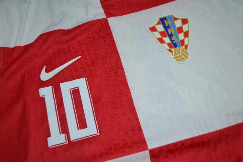 May include: A red and white checkered sports jersey featuring the number 10 in white, a white Nike swoosh logo, and the Croatian Football Federation crest with the letters HNS and a soccer ball graphic on the upper right chest area.