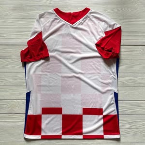 Puede incluir: Una camiseta deportiva de manga corta con un patr&oacute;n de cuadros blancos y rojos en la parte inferior, mangas rojas lisas, cuerpo superior blanco, cuello en V rojo y paneles verticales azules a lo largo de los costados del torso.