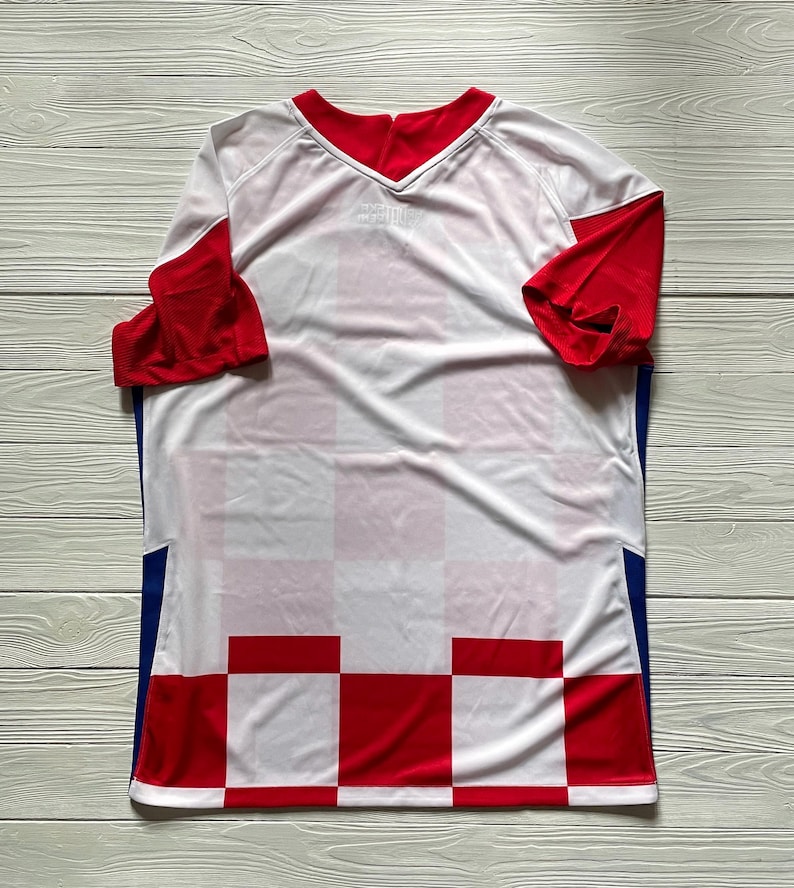 Puede incluir: Una camiseta deportiva de manga corta con un patr&oacute;n de cuadros blancos y rojos en la parte inferior, mangas rojas lisas, cuerpo superior blanco, cuello en V rojo y paneles verticales azules a lo largo de los costados del torso.
