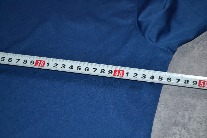 May include: A navy blue fabric is measured with a white and red ruler. The ruler shows measurements from 5 to 50 inches, equivalent to approximately 12.7 to 127 cm. The fabric is smooth.