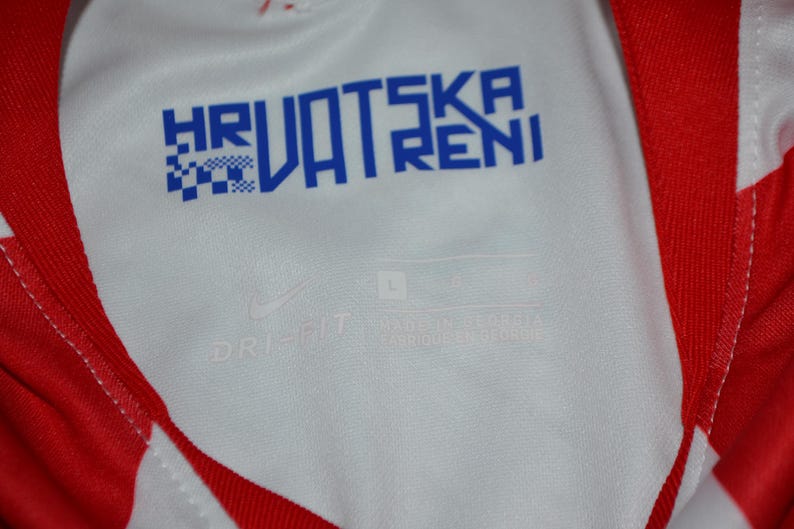 Puede incluir: Una camiseta deportiva blanca y roja con el texto azul HRVATSKA VATRENI impreso en el cuello interior. La tela incluye un logotipo de Nike y el texto DRI-FIT, con una etiqueta de talla L visible en la zona interior del cuello de la prenda.