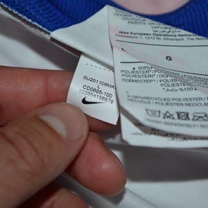 Puede incluir: Una prenda blanca con cuello azul que presenta una etiqueta de cuidado interna. La etiqueta muestra el logotipo de Nike, el texto SU201106MA, CD0695-100, 103654156879 e indica 100% poli&eacute;ster reciclado en talla grande con varios s&iacute;mbolos de cuidado.