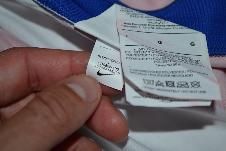 Puede incluir: Una prenda blanca con cuello azul que presenta una etiqueta de cuidado interna. La etiqueta muestra el logotipo de Nike, el texto SU201106MA, CD0695-100, 103654156879 e indica 100% poli&eacute;ster reciclado en talla grande con varios s&iacute;mbolos de cuidado.