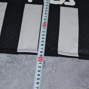 Puede incluir: Tela a rayas blancas y negras con el nombre de la marca "Fila" en blanco. Una cinta m&eacute;trica se coloca verticalmente a lo largo de la tela, mostrando medidas en cent&iacute;metros.